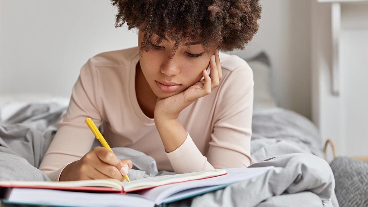 Image of beautiful woman with Afro hairstyle rewrites information in notebook from book, writes down homework, enjoys leisure time in bedroom, wears casual clothes. Female makes future plans