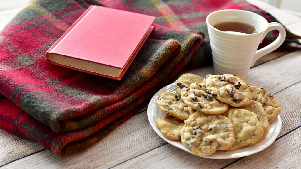 Photo of a plate of peanut butter chocolate chunk cookies with a book and a cup of tea